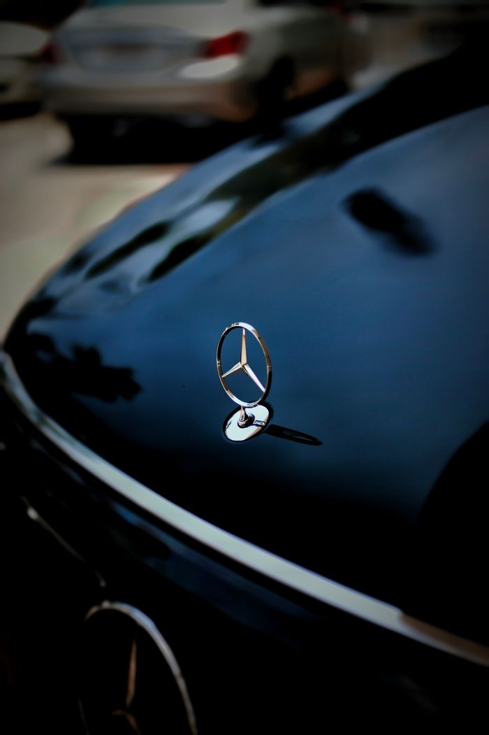 contact-img Detailed close-up of a car hood emblem reflecting on a shiny surface with blurred vehicles in the background.